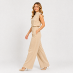 A woman modeling a beige sleeveless jumpsuit with a mock neck and wide-leg pants, paired with beige high heels, against a plain light gray studio background.