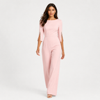 A woman modeling a light pink jumpsuit with three-quarter sleeves and decorative gold studs on the cuffs, set against a plain white studio background.
