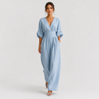 A woman modeling a light blue wide-leg jumpsuit with a V-neckline, elbow-length sleeves, and a matching fabric belt at the waist, photographed against a plain light gray studio background.