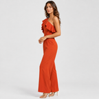 A woman modeling a rust-orange one-shoulder ruffle jumpsuit with wide-leg pants, paired with clear high-heeled sandals, against a light gray studio background.