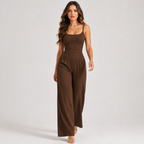 A woman modeling a sleeveless, high-waisted brown jumpsuit with wide legs, photographed against a plain light gray studio background.