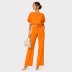 A woman modeling a bright orange short-sleeve jumpsuit with a tie waist, paired with beige heels and holding a beige handbag, against a light gray studio background.
