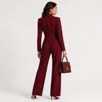 A woman stands with her back to the camera wearing a deep burgundy wide-leg jumpsuit and holding a matching burgundy handbag against a plain light gray studio background.