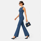 A woman models a sleeveless blue denim jumpsuit with wide-leg flared pants, walking in profile against a light gray studio background. She is holding a small navy blue handbag with gold hardware and wearing dark sunglasses and black heels.