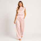 A woman modeling a light pink satin two-piece set consisting of a sleeveless mock-neck top and matching wide-leg trousers, photographed against a clean white studio background.