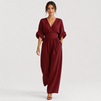 A woman modeling a burgundy wrap-style jumpsuit with elbow-length puff sleeves, a gathered waist, and wide legs, photographed against a clean light gray studio background.