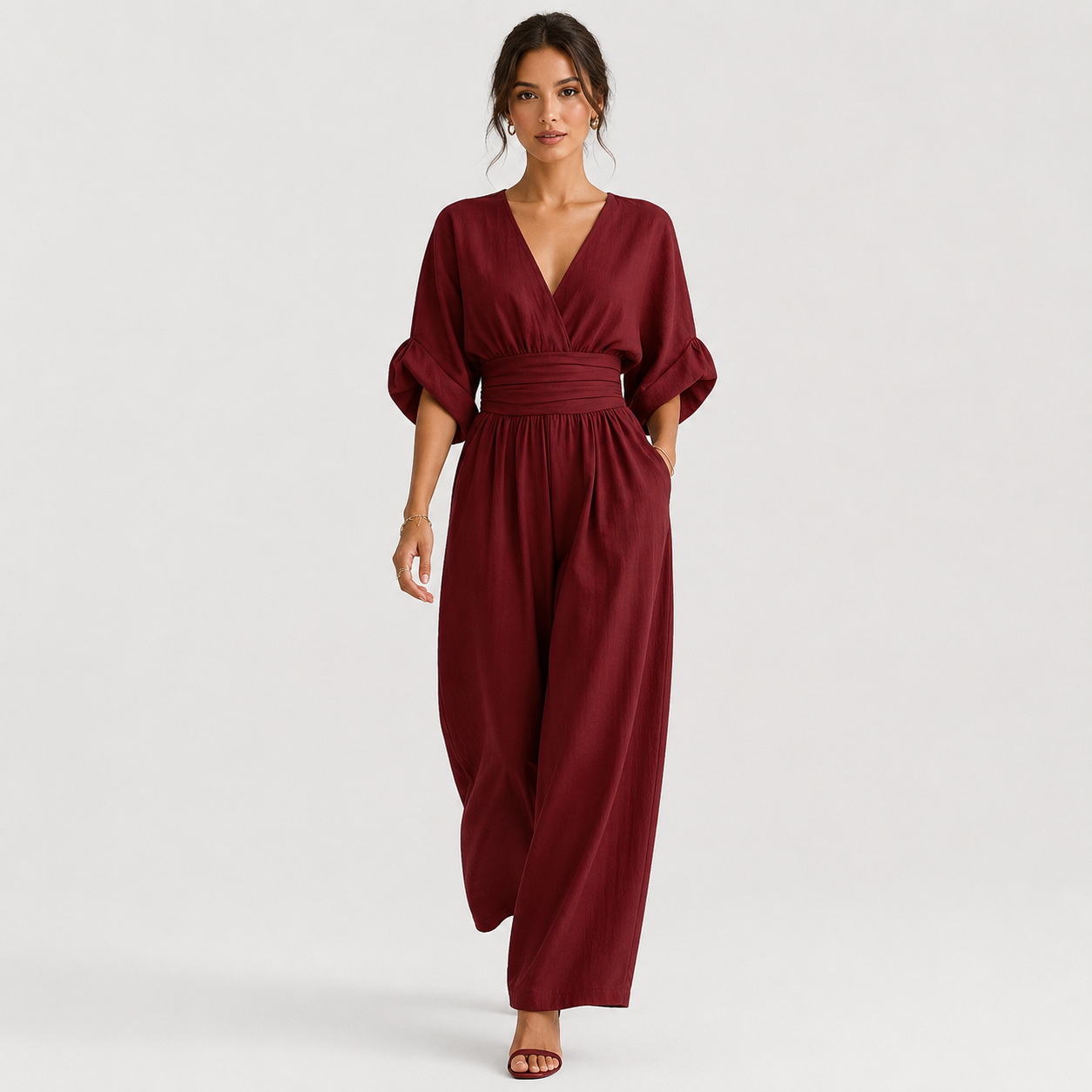 A woman modeling a burgundy wrap-style jumpsuit with elbow-length puff sleeves, a gathered waist, and wide legs, photographed against a clean light gray studio background.