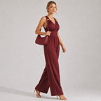 A woman models a sleeveless burgundy jumpsuit with a V-neckline, crossover shoulder straps, a matching fabric belt at the waist, and wide-leg pants. She is carrying a matching burgundy shoulder bag and wearing nude high-heeled sandals against a neutral gray studio background.