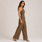 A woman modeling a one-shoulder wide-leg jumpsuit in a solid brown color, standing against a neutral light gray studio background.