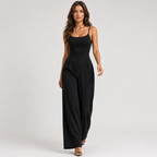 A woman modeling a black sleeveless jumpsuit with thin spaghetti straps, a square neckline, a defined waist, and wide-leg pants, photographed against a light gray studio background.
