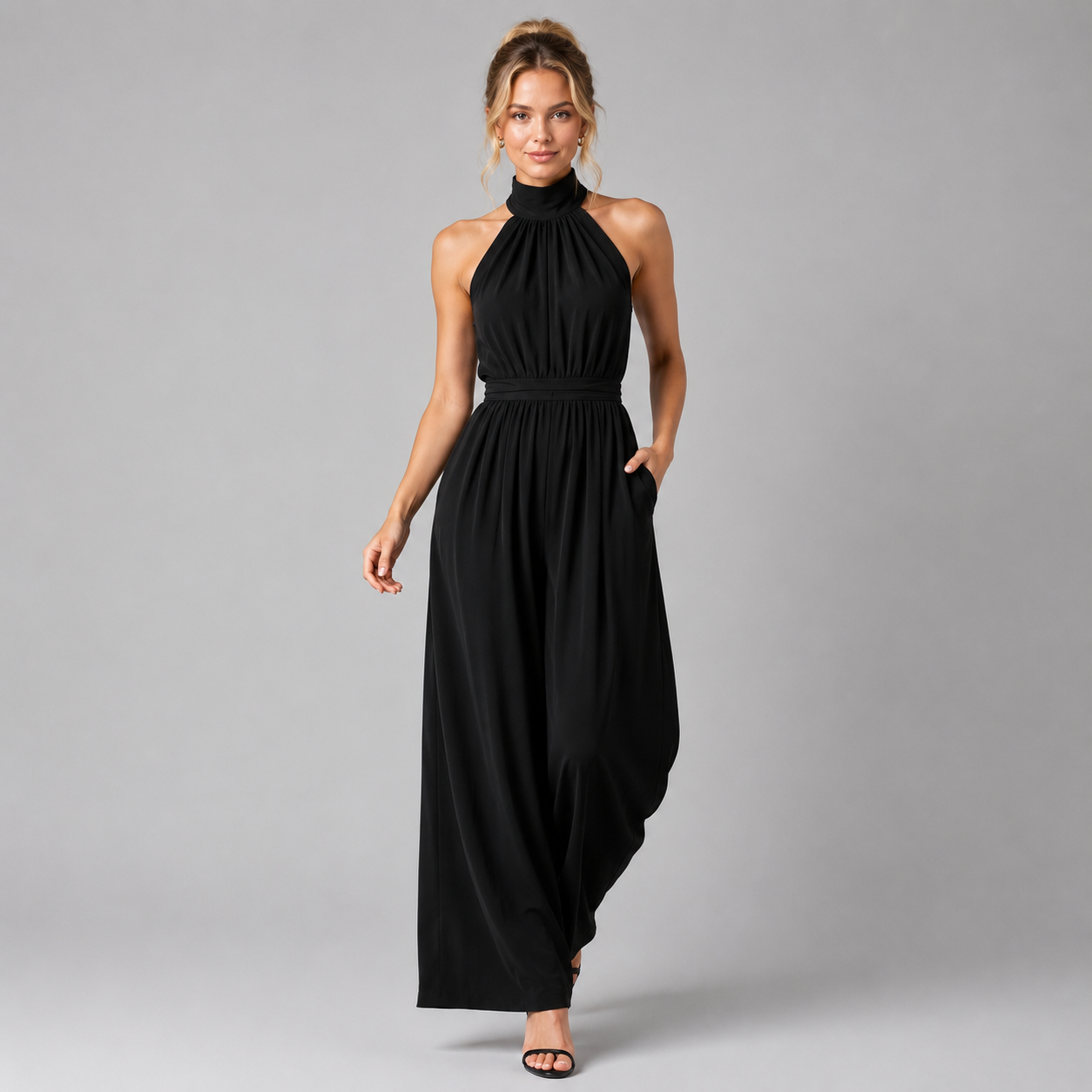 A woman modeling a black halter-neck maxi dress with side pockets, standing against a solid gray studio background.