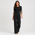 A woman modeling a black three-quarter sleeve jumpsuit with pearl embellishments on the sleeve cuffs, photographed against a light gray studio background.