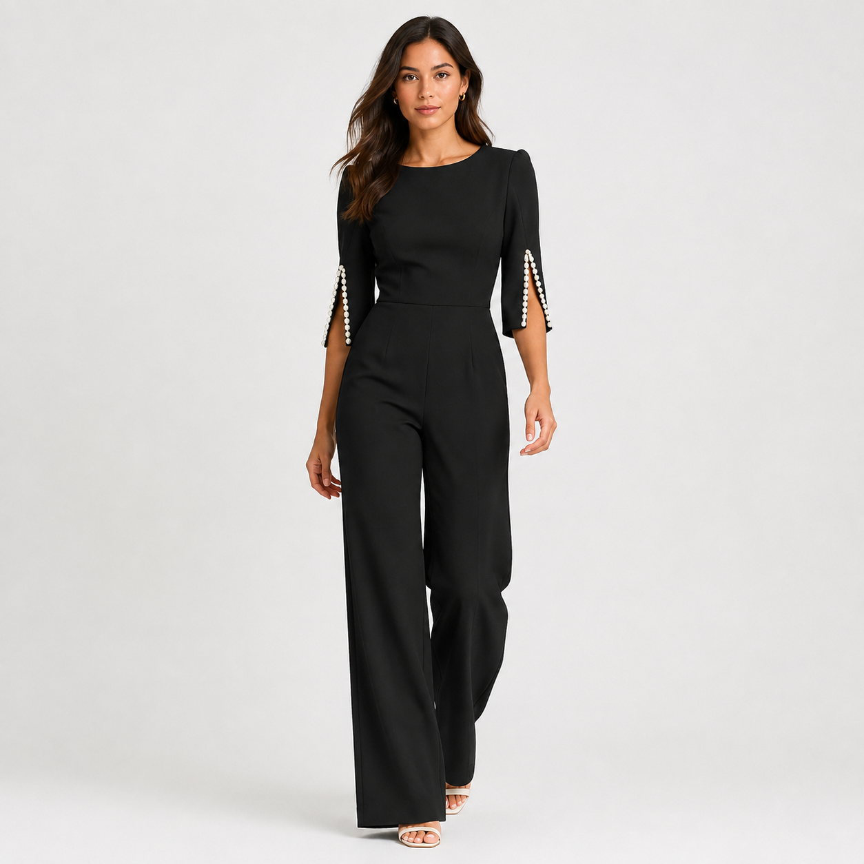 A woman modeling a black three-quarter sleeve jumpsuit with pearl embellishments on the sleeve cuffs, photographed against a light gray studio background.