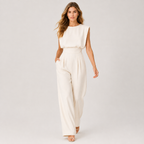 A woman modeling a sleeveless cream-colored jumpsuit with a high-waisted, wide-leg design, set against a plain light gray studio background.