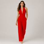 A woman modeling a bright red sleeveless halter jumpsuit with a deep V-neckline and wide-leg pants, photographed against a neutral light gray studio background.
