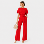 A woman modeling a bright red short-sleeve jumpsuit with a tie waist and wide-leg pants, holding a beige handbag against a plain light gray studio background.