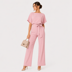 A woman modeling a dusty pink two-piece set consisting of a short-sleeve top and matching wide-leg trousers with a tie waist, holding a beige handbag against a plain white background.
