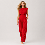 A woman modeling a bright red sleeveless jumpsuit with a high-waisted, wide-leg design, photographed against a neutral light gray studio background.