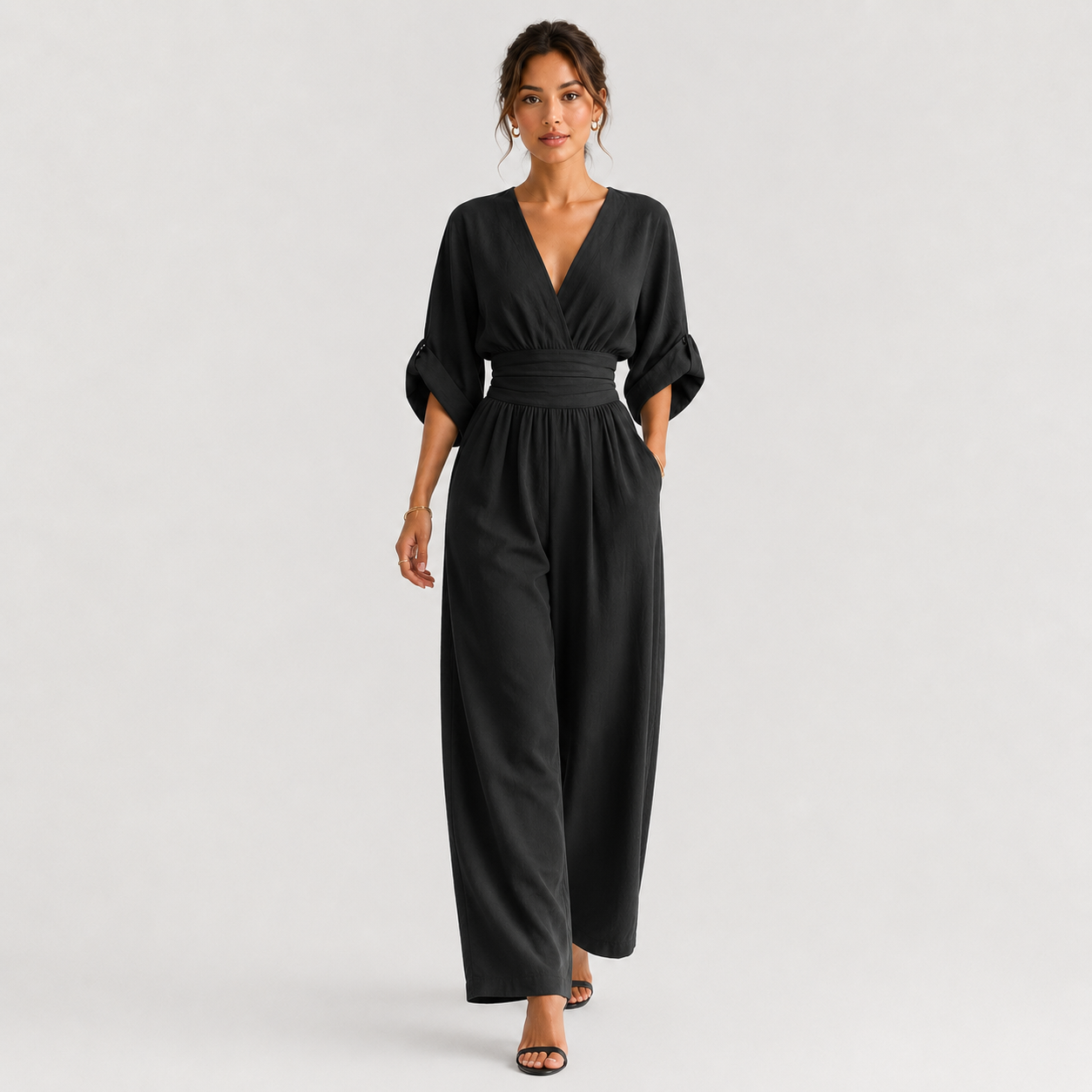 A woman modeling a black wide-leg jumpsuit with a wrap-style V-neck bodice, three-quarter sleeves, and a matching waistband, photographed against a clean light gray studio background.
