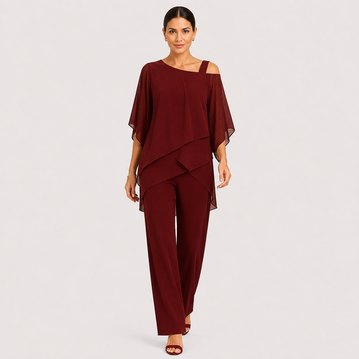 A woman modeling a burgundy two-piece outfit consisting of a cold-shoulder, asymmetrical draped top and matching wide-leg pants, photographed against a light gray studio background.