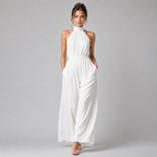 A woman modeling a white sleeveless halter-neck jumpsuit with wide legs, standing against a neutral gray studio background.