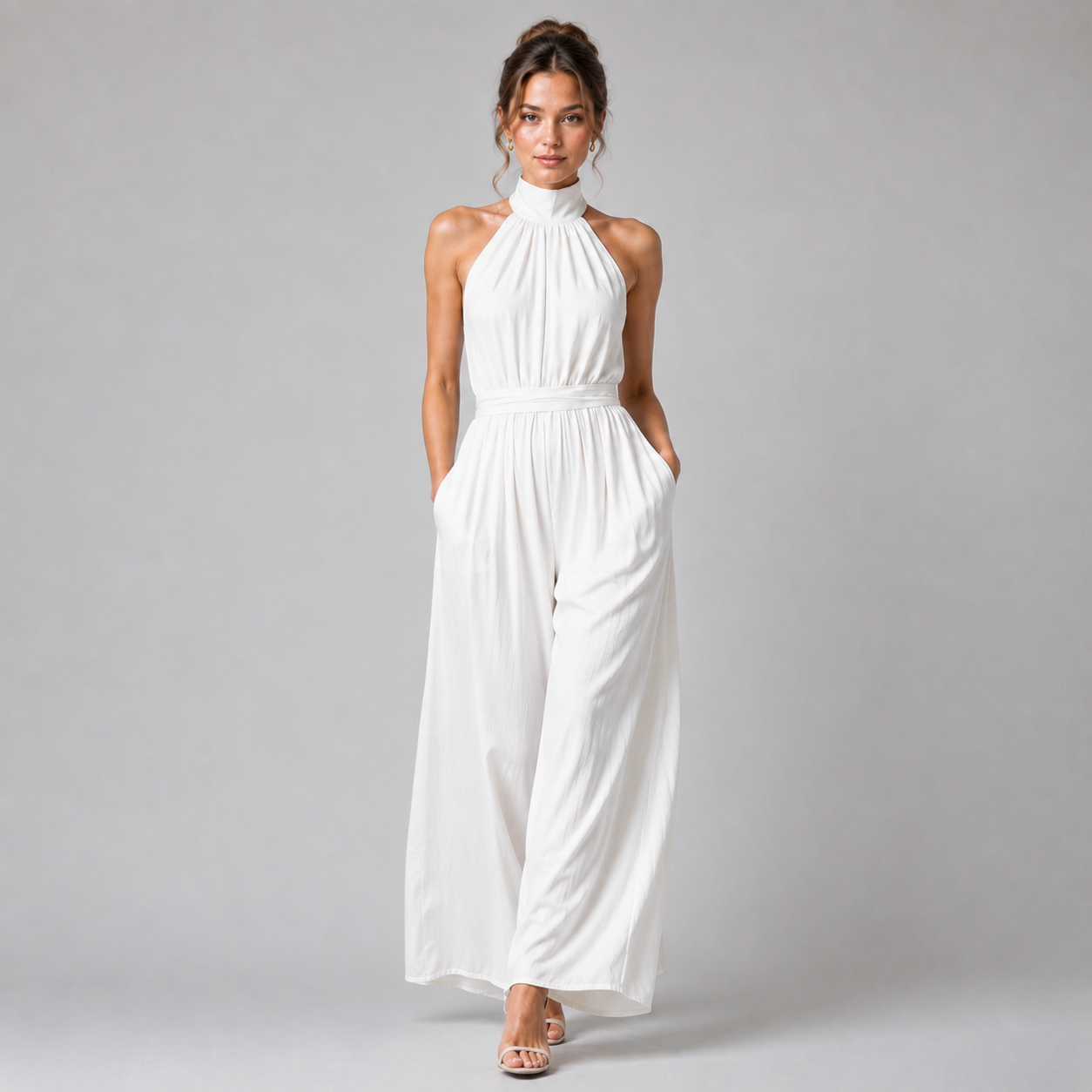 A woman modeling a white sleeveless halter-neck jumpsuit with wide legs, standing against a neutral gray studio background.