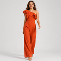 A woman modeling a bright rust-orange one-shoulder jumpsuit with dramatic ruffles across the neckline and wide legs, photographed against a clean light gray studio background.