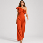 A woman modeling a bright rust-orange one-shoulder jumpsuit with dramatic ruffles across the neckline and wide legs, photographed against a clean light gray studio background.