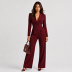 A woman modeling a deep burgundy wrap-style jumpsuit with long sleeves and wide legs, holding a matching burgundy handbag against a light gray studio background.