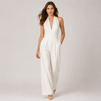 A woman modeling a white halter-neck jumpsuit with a deep V-neckline, defined waist seam, and wide-leg pants, set against a plain light gray studio background.