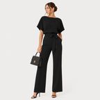 A woman modeling a black short-sleeve jumpsuit with a tie waist, paired with black high heels and holding a black handbag, against a light gray studio background.