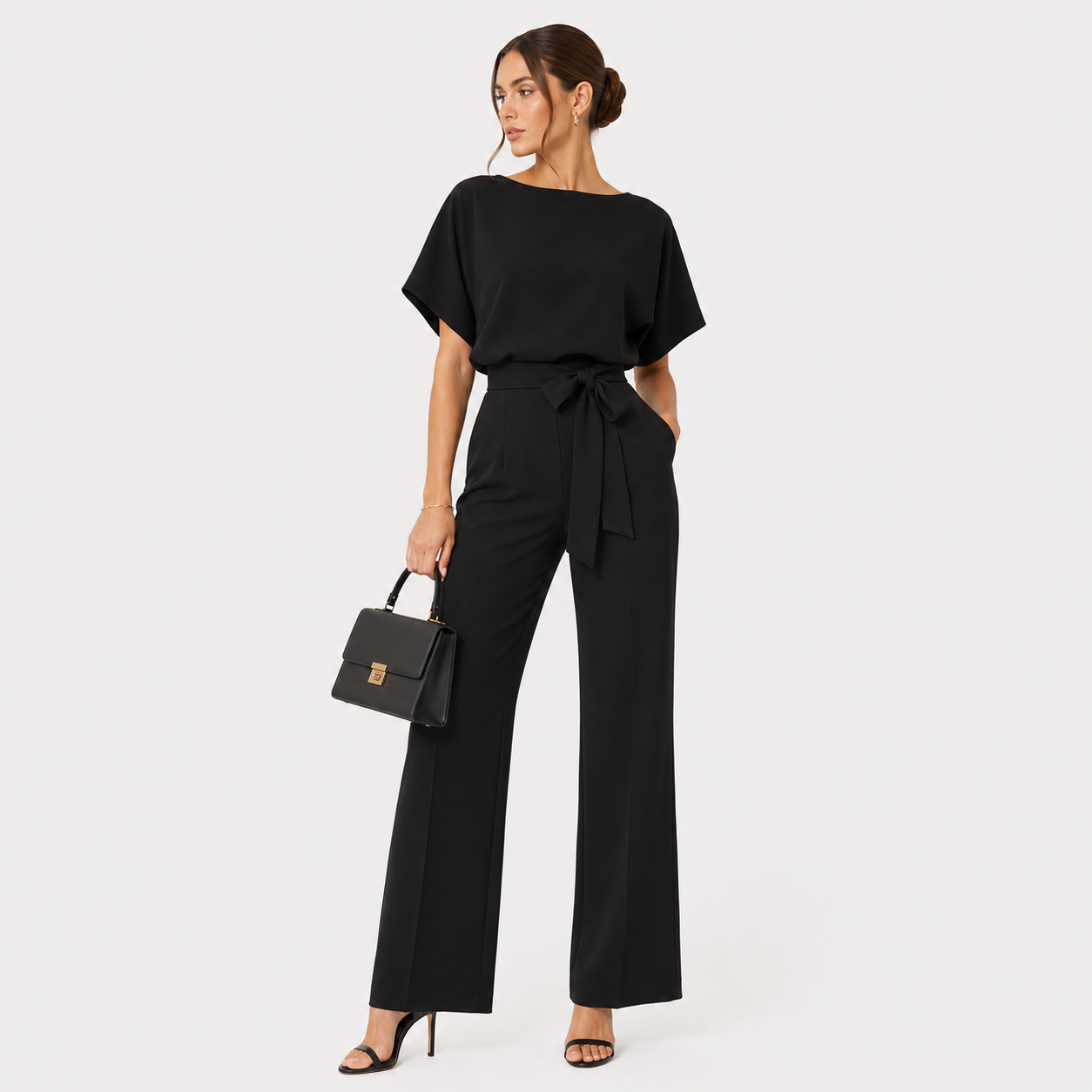 A woman modeling a black short-sleeve jumpsuit with a tie waist, paired with black high heels and holding a black handbag, against a light gray studio background.