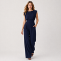 A woman modeling a navy blue sleeveless jumpsuit with a boat neckline, defined waist, and wide-leg pants, standing against a light gray studio background.