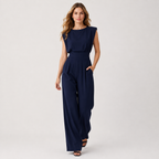 A woman modeling a navy blue sleeveless jumpsuit with a boat neckline, defined waist, and wide-leg pants, standing against a light gray studio background.