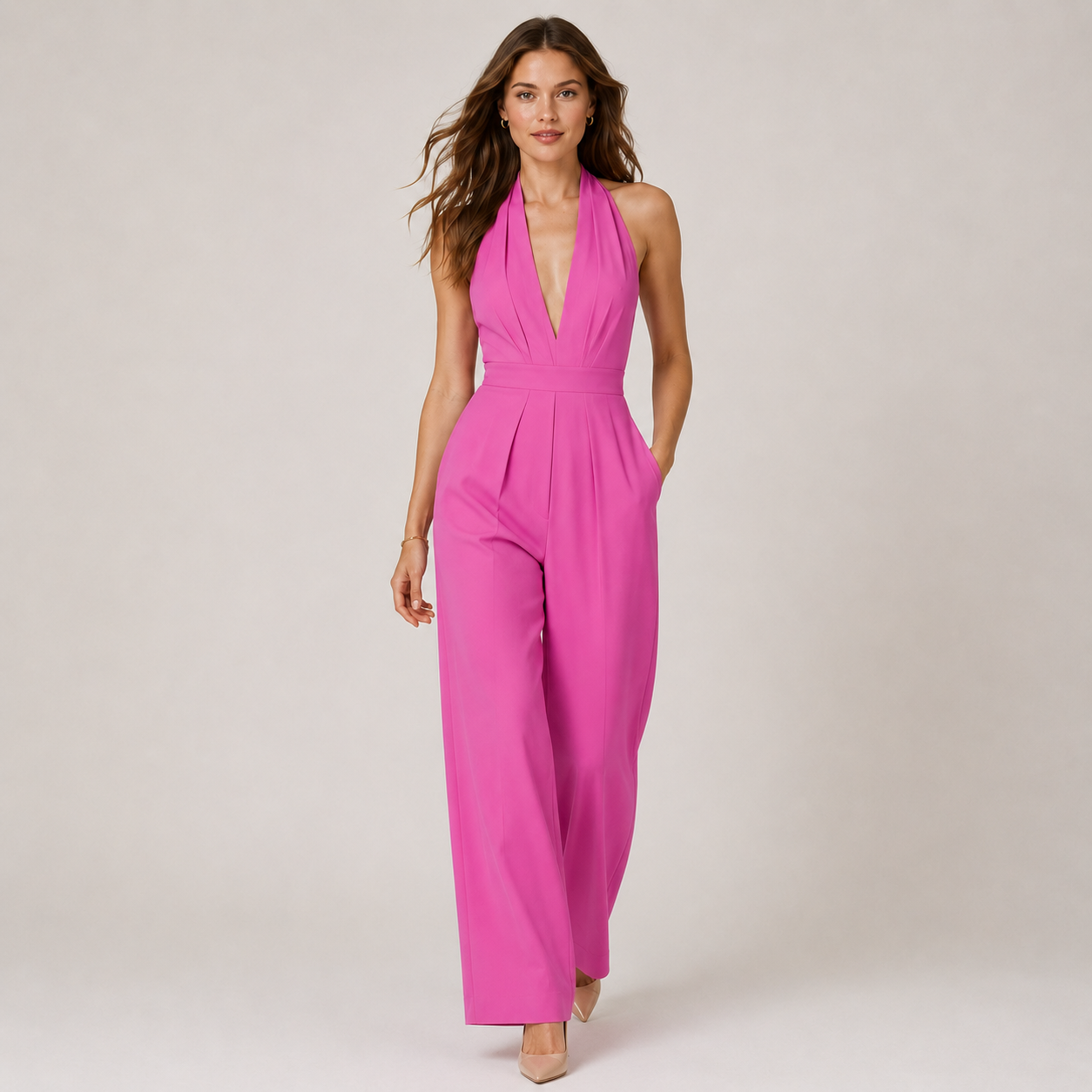 A woman modeling a bright pink halter-neck jumpsuit with a deep V-neckline and wide-leg pants, photographed against a neutral light beige studio background.