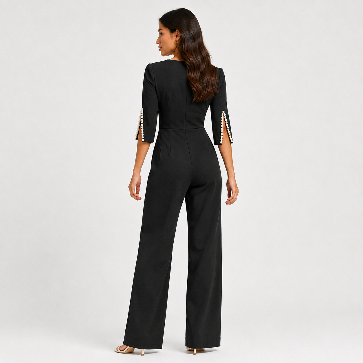 A woman viewed from the back wearing a black wide-leg jumpsuit with three-quarter sleeves and decorative pearl-like embellishments on the sleeve cuffs, photographed against a light gray studio background.