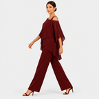 A woman modeling a burgundy two-piece outfit consisting of a cold-shoulder, tiered overlay top and matching wide-leg trousers, paired with dark heeled sandals, against a light gray studio background.