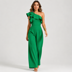 A woman modeling a bright green one-shoulder jumpsuit with a dramatic ruffled neckline and wide-leg pants, photographed against a clean light gray studio background.