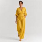 A woman modeling a mustard yellow wrap-style jumpsuit with elbow-length puff sleeves, a gathered waist, and wide legs, photographed against a light gray studio background.