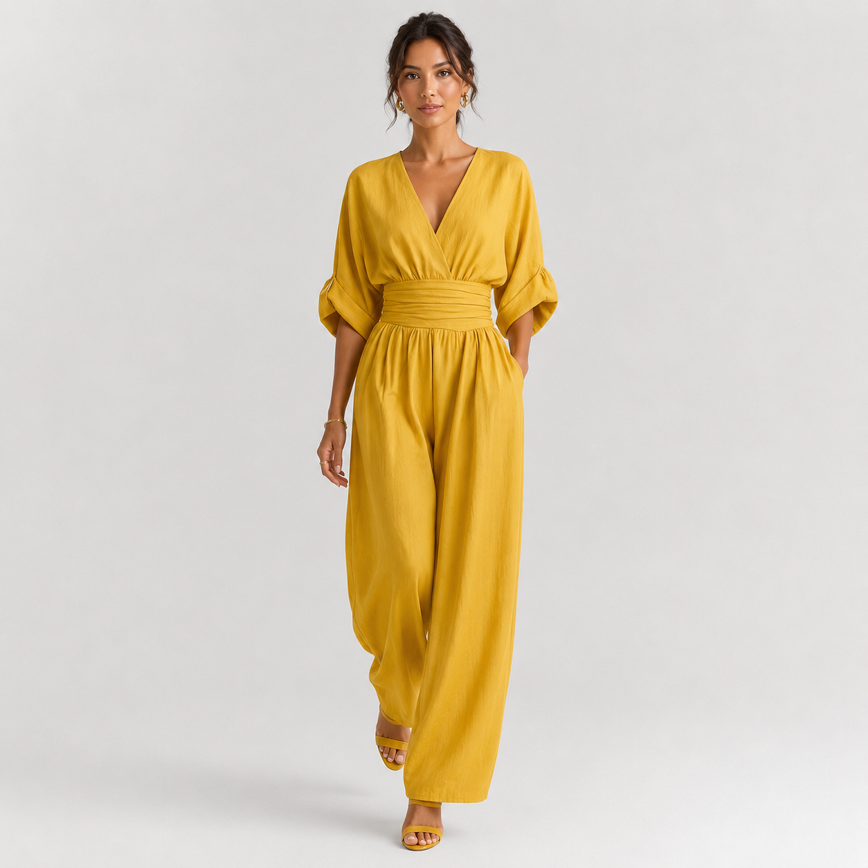 A woman modeling a mustard yellow wrap-style jumpsuit with elbow-length puff sleeves, a gathered waist, and wide legs, photographed against a light gray studio background.
