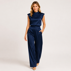 A woman modeling a navy blue satin jumpsuit with a mock neck and cap sleeves, set against a plain light gray studio background.