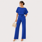 A woman models a royal blue two-piece jumpsuit featuring a short flutter-sleeve top and matching wide-leg trousers with a tie waist. She is holding a beige handbag and wearing beige high-heeled sandals, set against a solid light gray background.