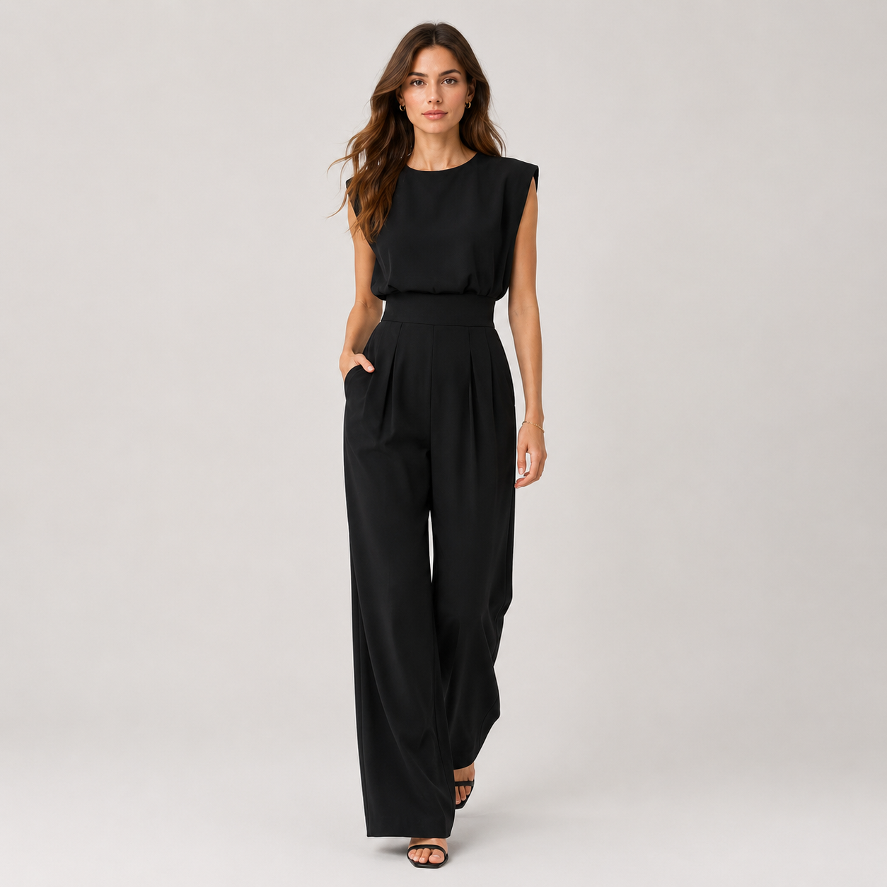 A woman modeling a black sleeveless jumpsuit with a high-waisted, wide-leg design, photographed against a neutral light gray studio background.