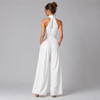 A woman viewed from the back wearing a white halter-neck wide-leg jumpsuit, standing against a plain gray studio background.