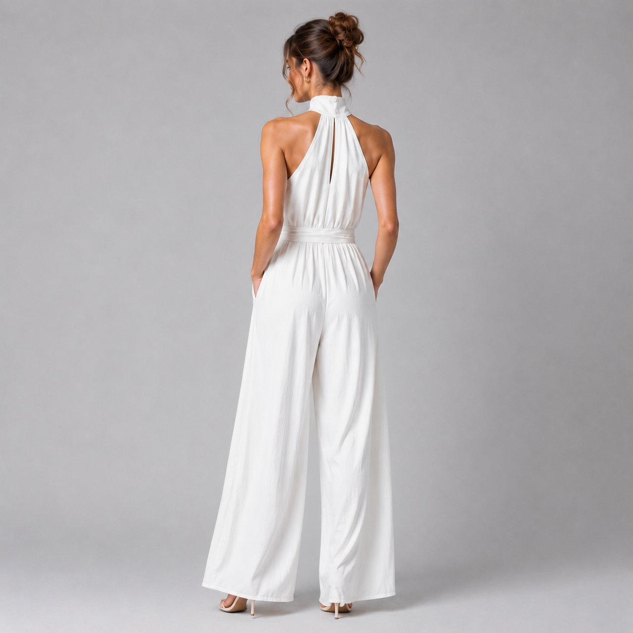 A woman viewed from the back wearing a white halter-neck wide-leg jumpsuit, standing against a plain gray studio background.
