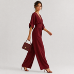 A woman models a deep burgundy wide-leg jumpsuit with three-quarter puff sleeves and a matching fabric belt at the waist. She is holding a matching burgundy handbag and wearing burgundy high-heeled sandals, set against a plain light gray studio background.