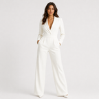 A woman modeling a white long-sleeve jumpsuit with a deep V-neckline, structured shoulders, and wide legs, standing against a light gray studio background.
