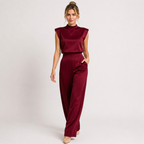 A woman modeling a two-piece satin jumpsuit in deep burgundy, featuring a sleeveless mock neck top and matching wide-leg trousers, set against a plain light gray studio background.