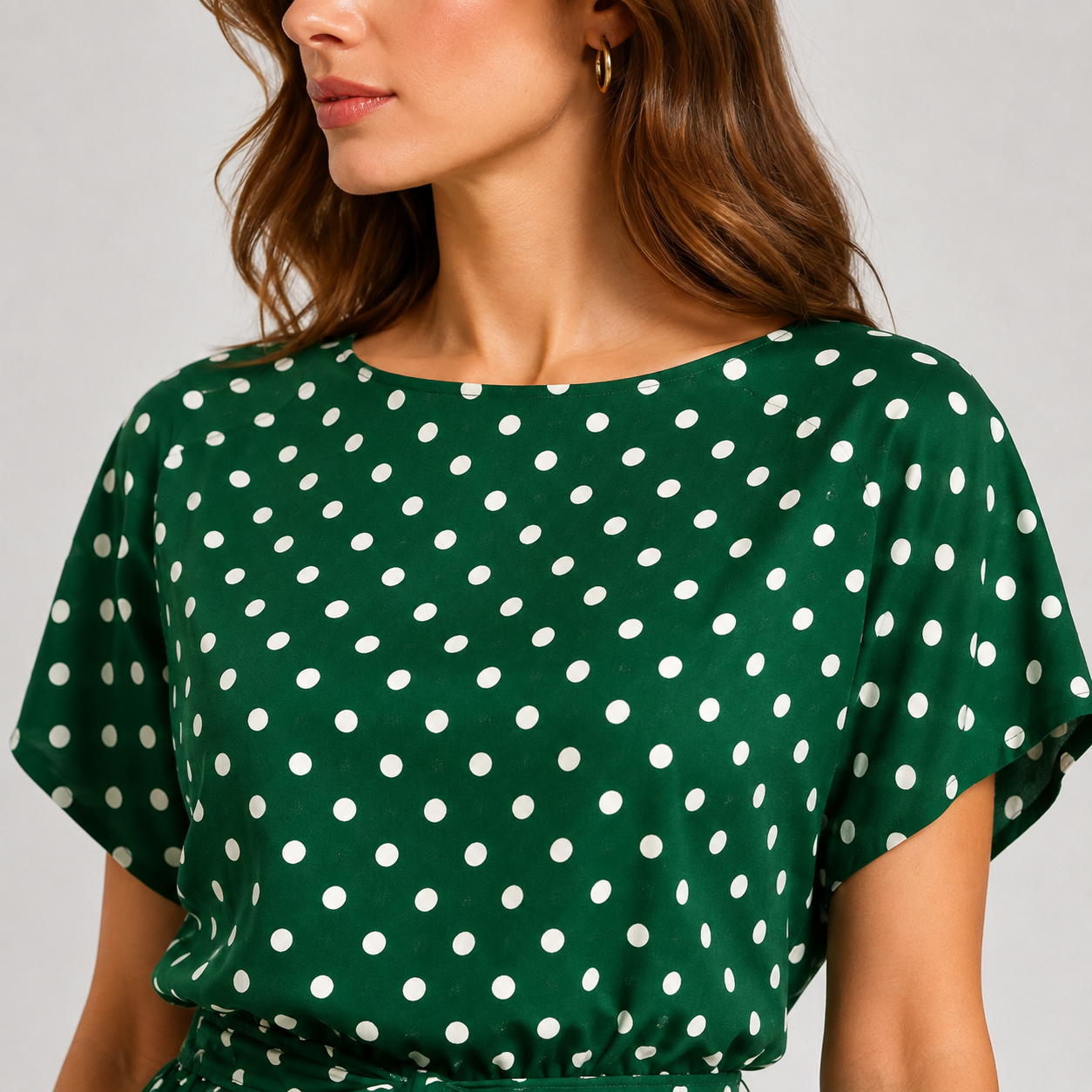A close-up of a woman wearing a green short-sleeve top with a white polka dot pattern, set against a plain light gray background.