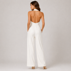 A woman shown from the back wearing a white sleeveless jumpsuit with a high open back, wide legs, and a halter-style neckline, photographed against a light gray studio background.